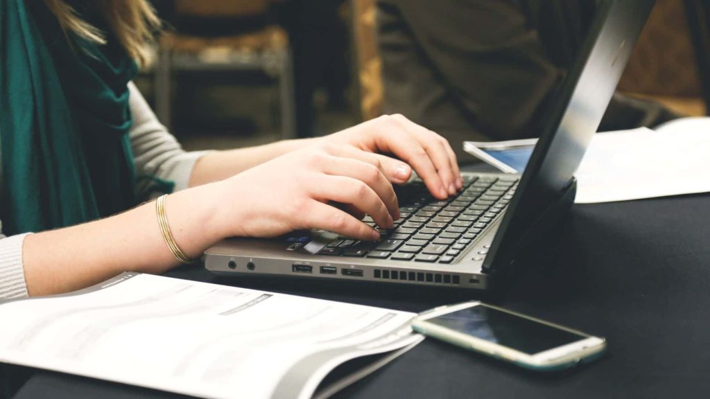 A person types on a laptop at a desk with documents and a smartphone. The scene conveys focus and professionalism in a work or study environment.