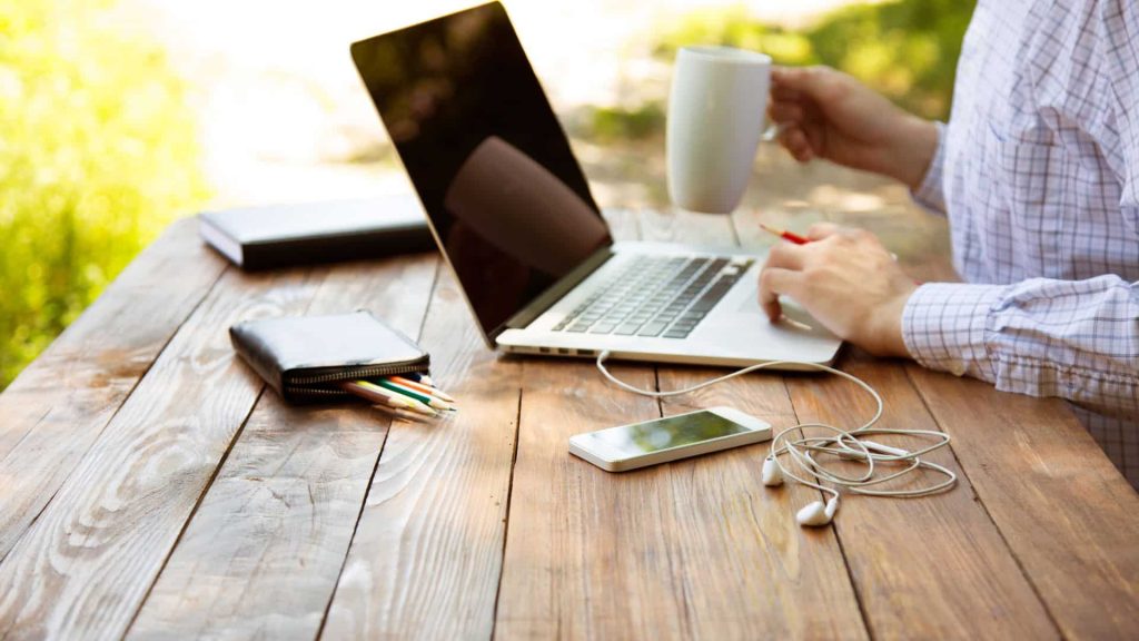 A person working outdoors on a wooden table with a laptop, holding a mug. Nearby are a smartphone, earphones, a notebook, and colored pencils. The scene conveys a relaxed, productive atmosphere.