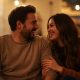 A couple sits on a couch, warmly lit, smiling at each other. The cozy indoor setting conveys a sense of warmth and intimacy.